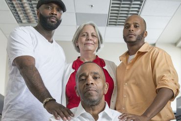 Darryl Pinkins, wearing a crisp, white button-up shirt, sits in a chair. He is surrounded by a few members of his support system: son, lawyer, nephew. His son and nephew each rest a hand on his shoulders.