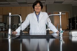 Hisako Masuda, wearing a white lab coat, stands in a chemistry lab behind a large, shiny black work table outfitted with silver instruments.