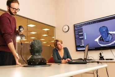 Miller and Johnson sit at a large table. While Johnson looks at the screen of a laptop, Miller holds a scanner pointed at a bust.