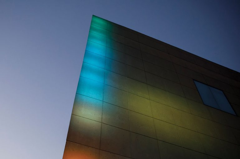 Multicolored lights shine on an angular stone facade that juts into the sky.