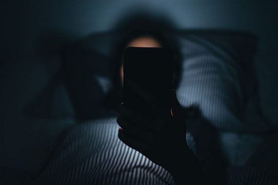 A person lays in her bed in a dark room, her face obscured by the back of a cell phone.