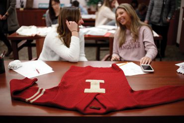 A red, vintage “I Man” sweater lays on a table in the foreground while two female students talk in the background.