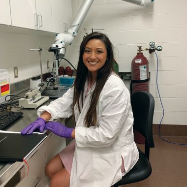 A woman with tan skin and long, black hair sits in a lab smiling. She is wearing a white lab coat and purple rubber gloves.