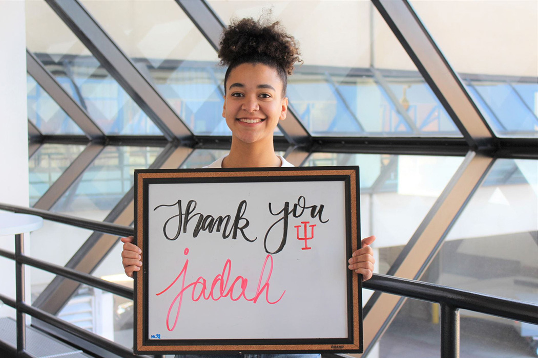 Jadah Cunningham, a woman with light-brown skin and curly hair in a ponytail on top of her head, smiles and holds a whiteboard on which she has written "Thank you! Jadah.”