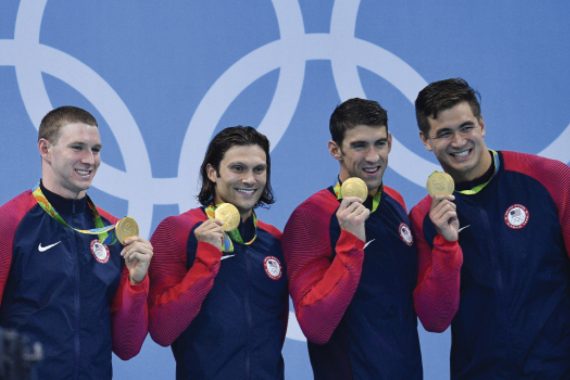 Four men wearing matching warm-up jackets stand shoulder to shoulder, smiling while holding up their Olympic gold medals.
