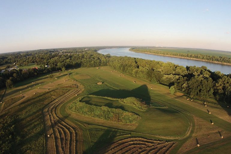 Aerial photograph of the Angel Mounds State Historic Site on a sunny day. At center is a large Earth mound surrounded by woodlands and bordered on the south by the winding Ohio River.