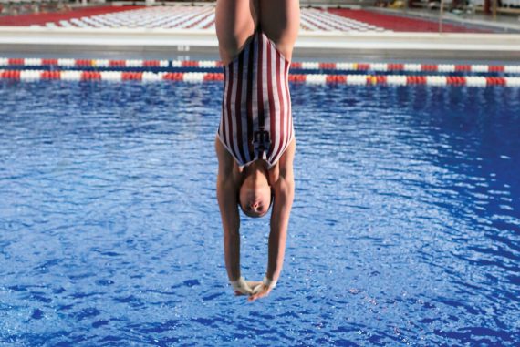 Photograph of a female diver, upside down in mid-dive, wearing a red-and-white striped IU swimsuit. The IU swimming pool and facility can be seen in the background.