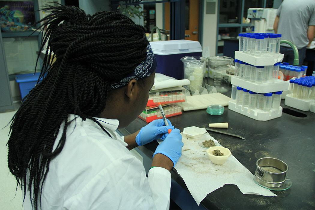 A woman wearing a white lab coat and blue gloves using a Sharpie while sitting at a lab table where small piles of dust and various containers sit.