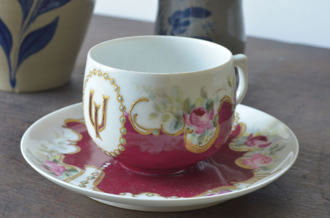 White teacup and saucer featuring magenta floral décor and an interlocking I and U (IU trident) embellished with gold accents.